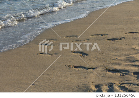 Footprints trailing along a serene beach as gentle waves caress the sandy shore at sunset. Footprints trailing along a serene beach as gentle waves caress the sandy shore at sunset. 133250456