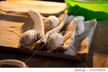 Raw bread dough loaves rising on linen cloth in baking tray, warm light highlighting texture, homemade organic food making process kitchen activity 133250926