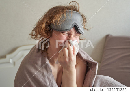 Sick caucasian female adult with sleep mask blowing nose in bed during illness 133251222