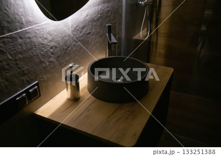 Stylish stone sink in a wooden countertop against the background of a mirror with diode lighting and a chrome tap. Fashionable interior, close-up Stylish stone sink in a wooden countertop against the background of a mirror with diode lighting and a chrome tap. Fashionable interior, close-up 133251388