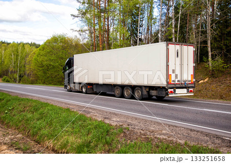 A truck with a refrigerated semi-trailer transports refrigerated cargo along a country road against the backdrop of beautiful nature and forest in summer. Copy space for text 133251658