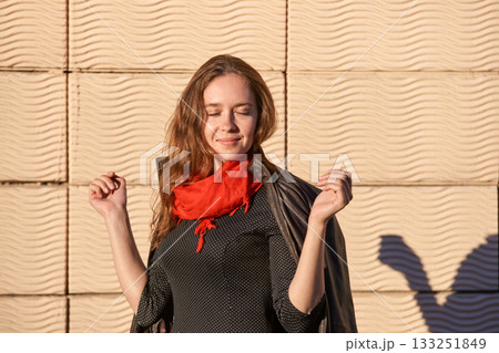Caucasian young female in red scarf enjoying sunlight against textured wall 133251849
