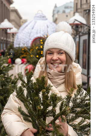 Smiling woman in a white knitted hat and beige coat enjoys festive winter decorations with Christmas trees and lights on a city street Smiling woman in a white knitted hat and beige coat enjoys festive winter decorations with Christmas trees and lights on a city street 133254934