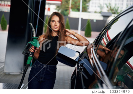 Leaning on the automobile, holding pump nozzle. A young woman at a gas station with her car 133254979