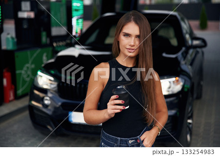 Black cup of the drink in hand. A young woman at a gas station with her car 133254983