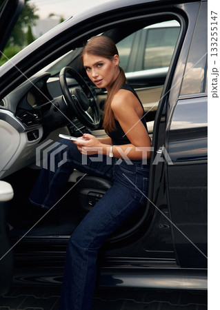 Looking at camera, holding smartphone. Woman is sitting in the car 133255147
