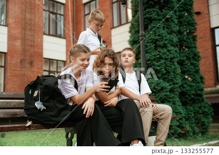 Sitting on the bench. Kids are near the school together, pupils 133255577