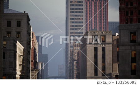 Tall buildings stretch towards the sky as the sun sets, casting a warm glow over the urban landscape. The streets below are quiet, creating a serene atmosphere in the city. 133256095