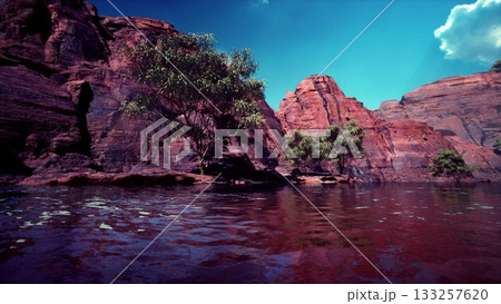 Majestic red rock cliffs rise above tranquil waters, creating a breathtaking landscape. Lush greenery contrasts against the rugged terrain under a clear blue sky. Natures beauty captivates. 133257620