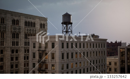 Dark clouds hang over urban buildings as the evening approaches. A water tower stands tall, while windows glow softly in the fading light, creating a serene urban atmosphere. Dark clouds hang over urban buildings as the evening approaches. A water tower stands tall, while windows glow softly in the fading light, creating a serene urban atmosphere. 133257642