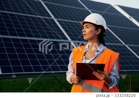 Black notepad in hands. Female worker engineer in uniform is near solar panels 133258220