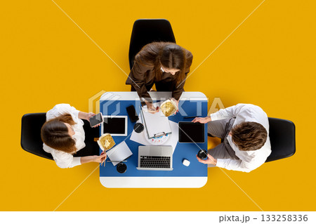 Office team eating take-away noodles while working with devices at blue desk. 133258336