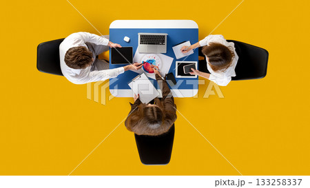 Team collaborating with digital devices around blue desk on yellow background in top-down view. Team collaborating with digital devices around blue desk on yellow background in top-down view. 133258337