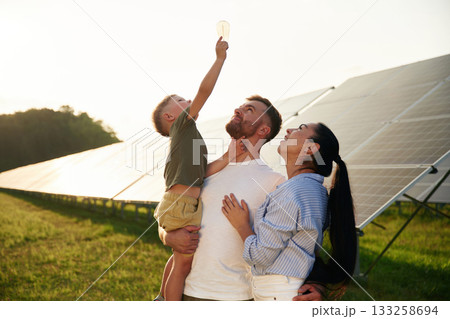 Light bulb in the hand. Father, mother and little son are outdoors near solar panels Light bulb in the hand. Father, mother and little son are outdoors near solar panels 133258694