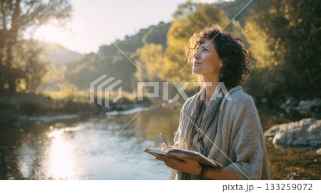 Inspirational Woman Taking Notes in a Journal by a Sunny Autumn River, Female Creative Writer or Artist Enjoying Solitude and Nature's Beauty at Sunset, Mindfulness, Wellness, and Travel Concept 133259072