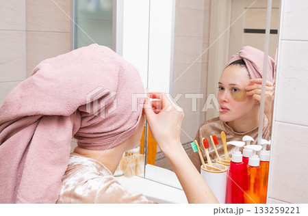 Woman in pink towel applying under eye patch for skin care in modern bathroom. 133259221