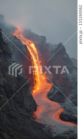 Vertical shot of a bright orange and red river of molten lava cascading down dark, rocky slope of an active volcano. The scene is dramatic and covered in mist or smoke, showcasing geology, raw nature Vertical shot of a bright orange and red river of molten lava cascading down dark, rocky slope of an active volcano. The scene is dramatic and covered in mist or smoke, showcasing geology, raw nature 133260062