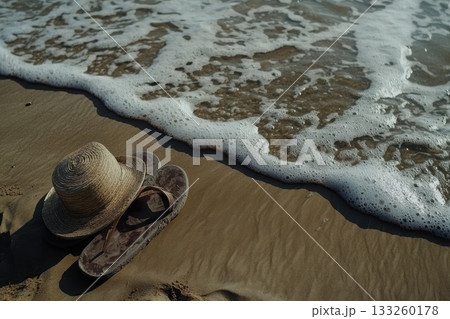 Close-up of a Pair of Flip Flops and a Sun Hat on a Sandy Beach Near the Ocean Waves 133260178