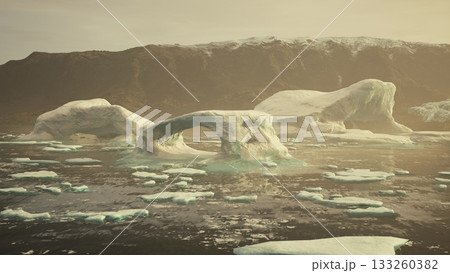 Icebergs with unique shapes drift peacefully in calm arctic waters as the sun sets, casting a warm glow over the icy landscape and rugged mountains in the background. 133260382