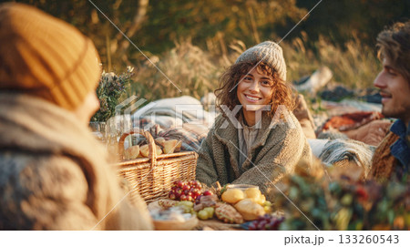 Smiling Young Woman in a Knitted Hat and Cozy Cardigan Enjoying an Autumn Picnic with Friends, Surrounded by Baskets, Food, and Blankets in the Warm Glow of Sunset 133260543