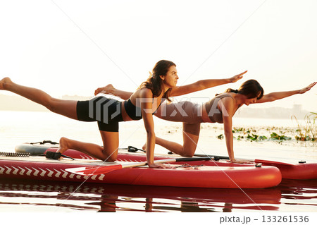 Doing yoga exercises, asana. Women friends are on sup board on the lake together Doing yoga exercises, asana. Women friends are on sup board on the lake together 133261536