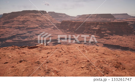 Vast red rock formations extend across the horizon, revealing intricate geological patterns. The sun illuminates the rugged terrain, creating a breathtaking view that draws exploration. 133261747