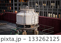 A large, white cooling tower stands prominently on a rooftop, surrounded by urban architecture. The twilight sky casts a warm glow on the city, highlighting the bustling energy below. 133262125