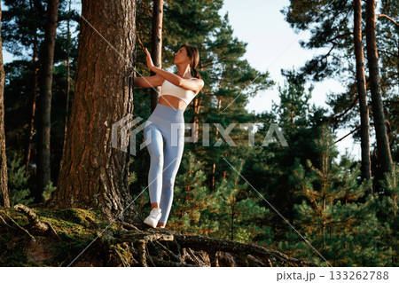 Holding by a tree. Woman in fitness clothes is in the forest Holding by a tree. Woman in fitness clothes is in the forest 133262788