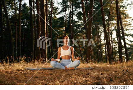 In blue pants and white top, sitting. Woman is doing yoga fitness exercises in the forest In blue pants and white top, sitting. Woman is doing yoga fitness exercises in the forest 133262894