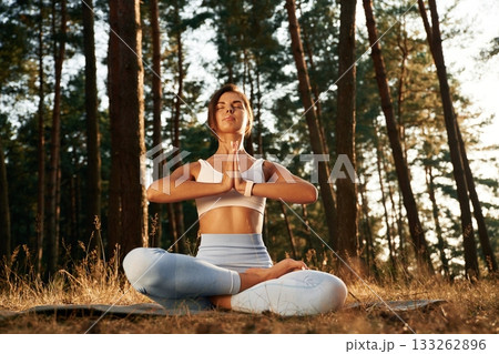 In blue pants and white top, sitting. Woman is doing yoga fitness exercises in the forest In blue pants and white top, sitting. Woman is doing yoga fitness exercises in the forest 133262896
