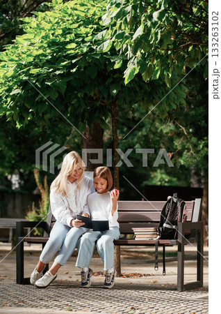 Learning and reading together. Little schoolgirl with her mother are sitting on the bench outdoors Learning and reading together. Little schoolgirl with her mother are sitting on the bench outdoors 133263102