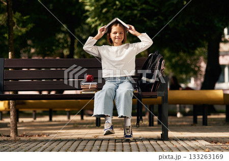 Having fun, book over the head. Schoolgirl is sitting on the bench with a bunch of books outdoors 133263169