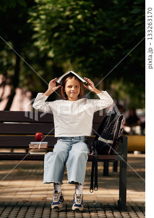 Having fun, book over the head. Schoolgirl is sitting on the bench with a bunch of books outdoors 133263170