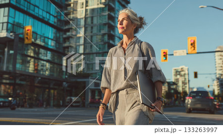 A confident mature businesswoman in a stylish striped outfit walks purposefully through a sunny city street, holding a laptop against a modern urban backdrop 133263997