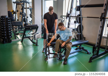 Professional sports trainer help disabled man in wheelchair move from exercise machine to wheelchair Professional sports trainer help disabled man in wheelchair move from exercise machine to wheelchair 133264008