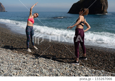 Women fitness stretching partners warm up on a sunny pebble beach by the ocean 133267066