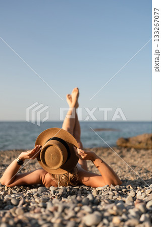 Beach summer relaxation. Woman lying on pebble beach covering face with straw hat, legs raised, enjoying sunny vacation day with copy space. Beach summer relaxation. Woman lying on pebble beach covering face with straw hat, legs raised, enjoying sunny vacation day with copy space. 133267077