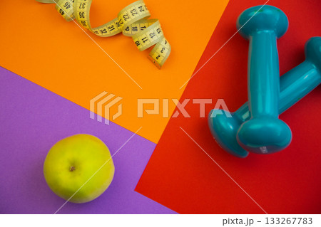 Vibrant flat lay arrangement with green apple, blue dumbbells, and measuring tape on colorful geometric backgrounds, symbolizing health, fitness, and wellness choices 133267783