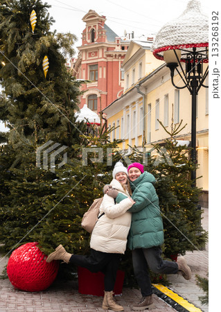 Two women in winter coats hug playfully among Christmas trees decorated with warm lights on a festive city street Two women in winter coats hug playfully among Christmas trees decorated with warm lights on a festive city street 133268192