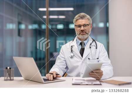 Professional senior male doctor wearing a white coat and stethoscope, sitting at a modern desk, working with a laptop and holding a digital tablet, providing healthcare and medical expertise 133268288