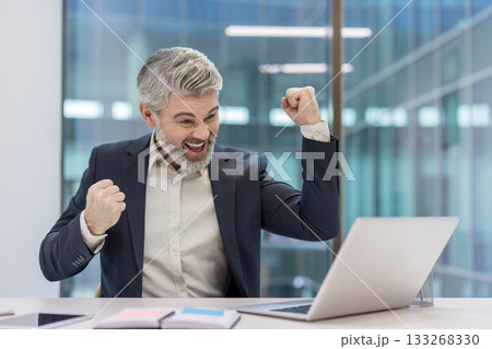 Excited mature businessman with a beard and gray hair smiling, raising fists in triumph while looking at a laptop screen in a modern office, celebrating business success or achievement 133268330