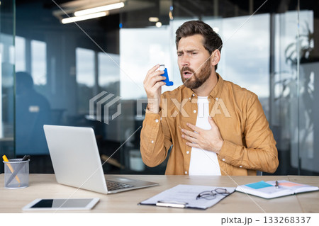 Man experiencing an asthma attack and breathing difficulty while using a blue preventative inhaler, holding hand on chest, sitting at his office desk with a laptop and documents Man experiencing an asthma attack and breathing difficulty while using a blue preventative inhaler, holding hand on chest, sitting at his office desk with a laptop and documents 133268337
