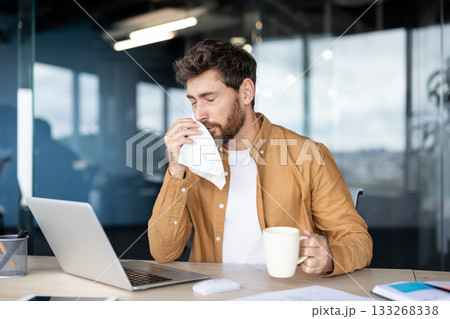 Man with a beard holding a tissue and blowing his nose while sitting at his desk, working on a laptop in a modern office, experiencing cold or flu symptoms Man with a beard holding a tissue and blowing his nose while sitting at his desk, working on a laptop in a modern office, experiencing cold or flu symptoms 133268338