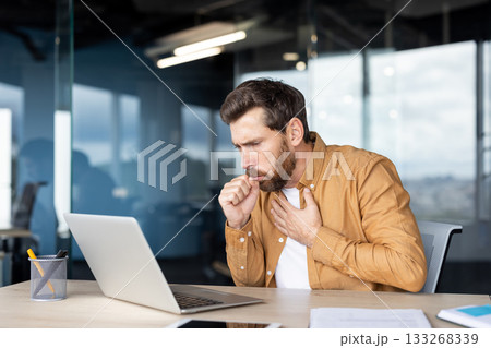 Businessman feeling unwell and coughing at his office desk, struggling to concentrate on laptop work while battling cold or flu symptoms in a modern corporate workspace 133268339