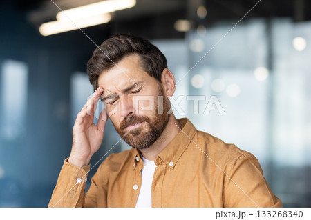 Tired man holding hand to his forehead, closing eyes from extreme headache and migraine pain, suffering from stress, mental health issues, and occupational burnout in an office environment Tired man holding hand to his forehead, closing eyes from extreme headache and migraine pain, suffering from stress, mental health issues, and occupational burnout in an office environment 133268340