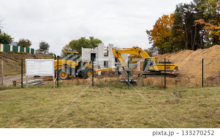 Yellow excavator and loader against the backdrop of a brick building under construction and a pile of sand. Construction equipment 133270252