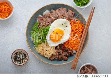Traditional korean bibimbap with fried egg, beef and vegetables and other toppings top view on light background Traditional korean bibimbap with fried egg, beef and vegetables and other toppings top view on light background 133270311