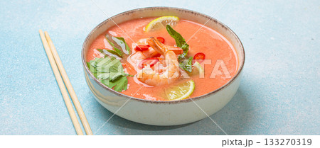 Traditional Thai soup tom yum soup with shrimp, lime, and herbs in bowl with chopsticks on blue background Traditional Thai soup tom yum soup with shrimp, lime, and herbs in bowl with chopsticks on blue background 133270319