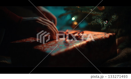 Close-up of hands tying a ribbon on a Christmas gift under a tree. The dark, cinematic scene creates a magical, intimate, and mysterious mood. Ideal for holiday greetings and festive ads. 133271493