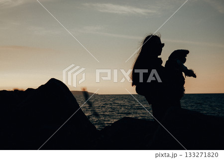 Silhouette of a woman holding a dog at sunset by the sea. A woman and her dog are silhouetted against the setting sun, with the ocean and a landscape 133271820